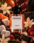 A bottle of L’AIMANT surrounded by orange slices, white flowers, vanilla beans, and sugar cubes, evoking a parfum sucré floral.