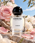 A glass bottle of CANDEUR Eau de Parfum with a black spherical cap, set on a stone surface surrounded by pink roses and white flowers, evoking a floral parfum.